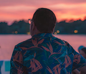 Back-view person in a blue tropical shirt and glasses watching a pink-orange lakeside sunset with soft bokeh lights