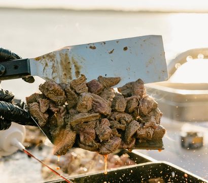 Sizzling grilled steak bites on a metal spatula, juices dripping onto a hot griddle during a sunny waterfront cookout.