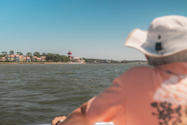 Person in a sun hat on a boat looking across choppy water toward a shoreline of pastel beach houses and a red-and-white striped lighthouse under a clear blue sky.