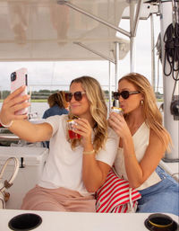 Two friends on a sailboat taking a selfie, wearing sunglasses and sipping cocktails with a striped beach bag and waterfront visible in the background.