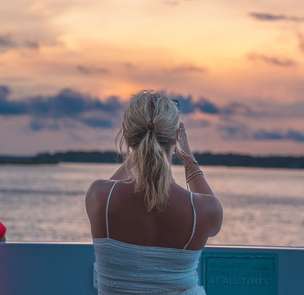 Back view of a person on a boat capturing a pastel coastal sunset over calm water, blonde hair in a ponytail and a silhouetted shoreline on the horizon.