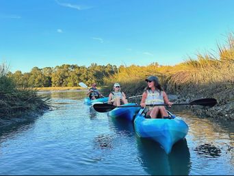 Three people in blue kayaks paddling a narrow coastal marsh channel at golden hour, surrounded by tall salt‑marsh grasses, calm water, and a clear blue sky.