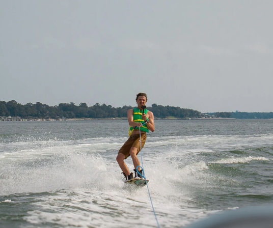 Person wakeboarding behind a boat on a tree-lined coastal inlet, wearing a green life jacket and brown shorts, splashing through the white wake