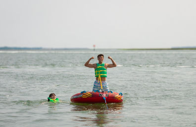 Two people in green life jackets enjoying water tubing on a red inflatable towable in a calm coastal inlet, one standing and flexing while the other swims nearby.