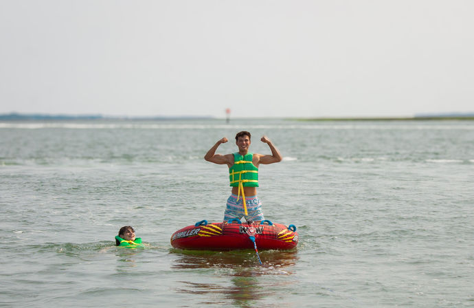 Two people in green life jackets enjoying water tubing on a red inflatable towable in a calm coastal inlet, one standing and flexing while the other swims nearby.
