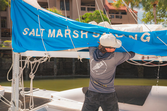 Person adjusting a bright blue sail cover with partial white lettering on a small sailboat at a sunny coastal marina, dock, ropes, and rigging visible.