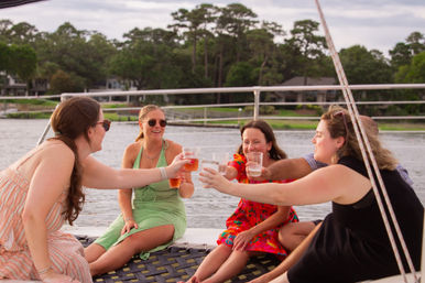 Group of friends toasting with drinks while seated on the netting of a sailboat, relaxing by a tree-lined waterfront on a warm afternoon.