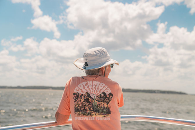 Sun‑hatted boater in a coral graphic tee leans on a boat railing, gazing across sunlit coastal waters under a blue sky dotted with puffy clouds.