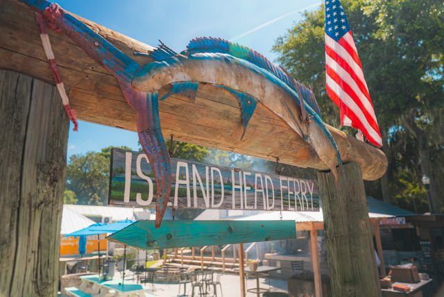 Rustic wooden ferry sign hung from a beam with a colorful metal fish sculpture above it, American flag waving and sunlit outdoor dock seating.