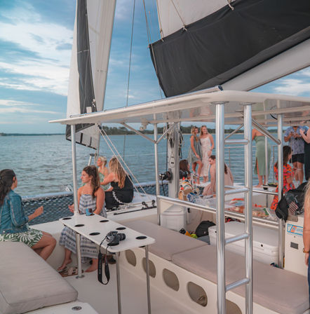 People socializing on a catamaran sailboat deck with white sails and metal ladder, camera on a table, cruising calm coastal waters under a partly cloudy sky — relaxed boat cruise vibe.