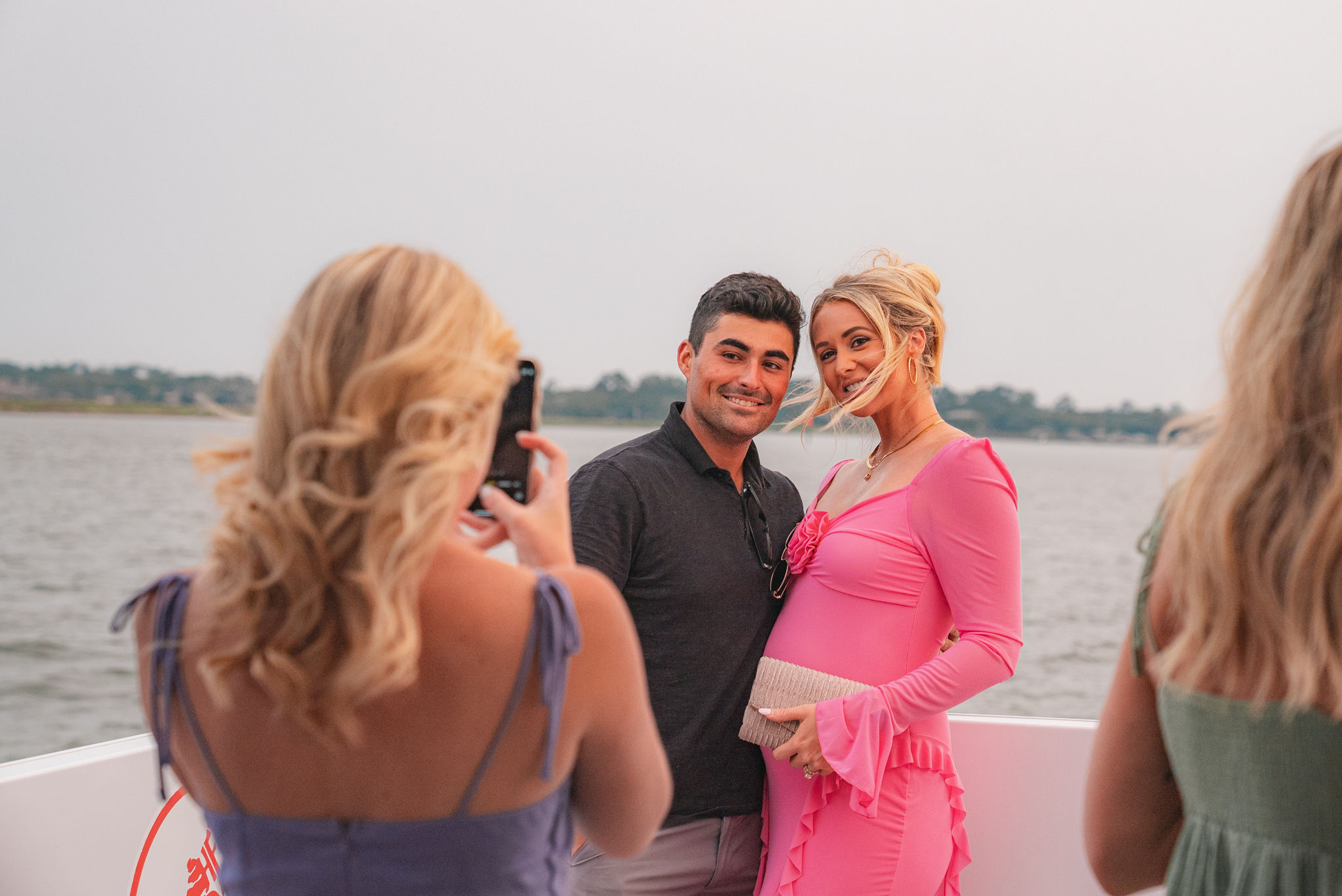 Couple posing on a boat by a calm shoreline at sunset — man in a dark shirt and woman in a bright pink dress cradling her belly while a friend snaps a smartphone photo.