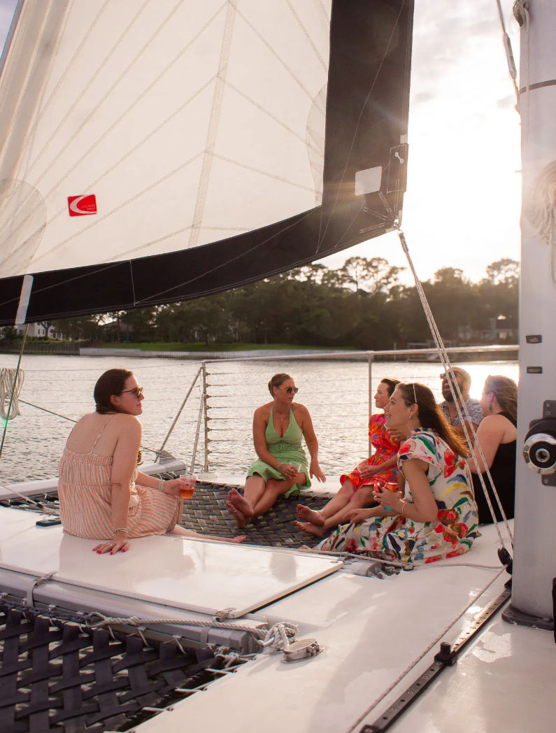 Group of friends relaxing on a catamaran trampoline under a large white sail during a sunset cruise on a calm coastal waterway, chatting and sipping drinks.