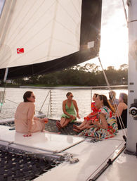 Group of friends relaxing on a catamaran trampoline under a large white sail during a sunset cruise on a calm coastal waterway, chatting and sipping drinks.