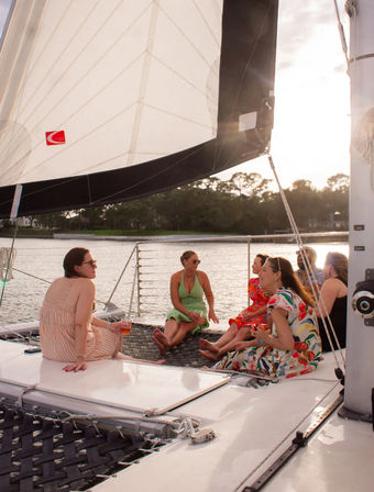 Group of friends relaxing on a catamaran trampoline under a large white sail during a sunset cruise on a calm coastal waterway, chatting and sipping drinks.