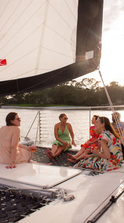 Group of friends relaxing on a catamaran trampoline under a large white sail during a sunset cruise on a calm coastal waterway, chatting and sipping drinks.