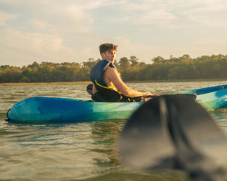 Young man wearing a blue life jacket paddling a teal kayak on a calm lake at golden hour, blurred paddle in the foreground and tree-lined shore in the background.