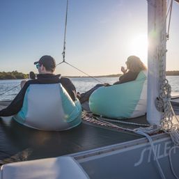 Two people lounging in mint-and-white beanbags on a sailboat deck at golden-hour sunset, sipping drinks and watching calm water and a tree-lined shore