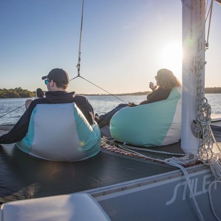 Two people lounging in mint-and-white beanbags on a sailboat deck at golden-hour sunset, sipping drinks and watching calm water and a tree-lined shore