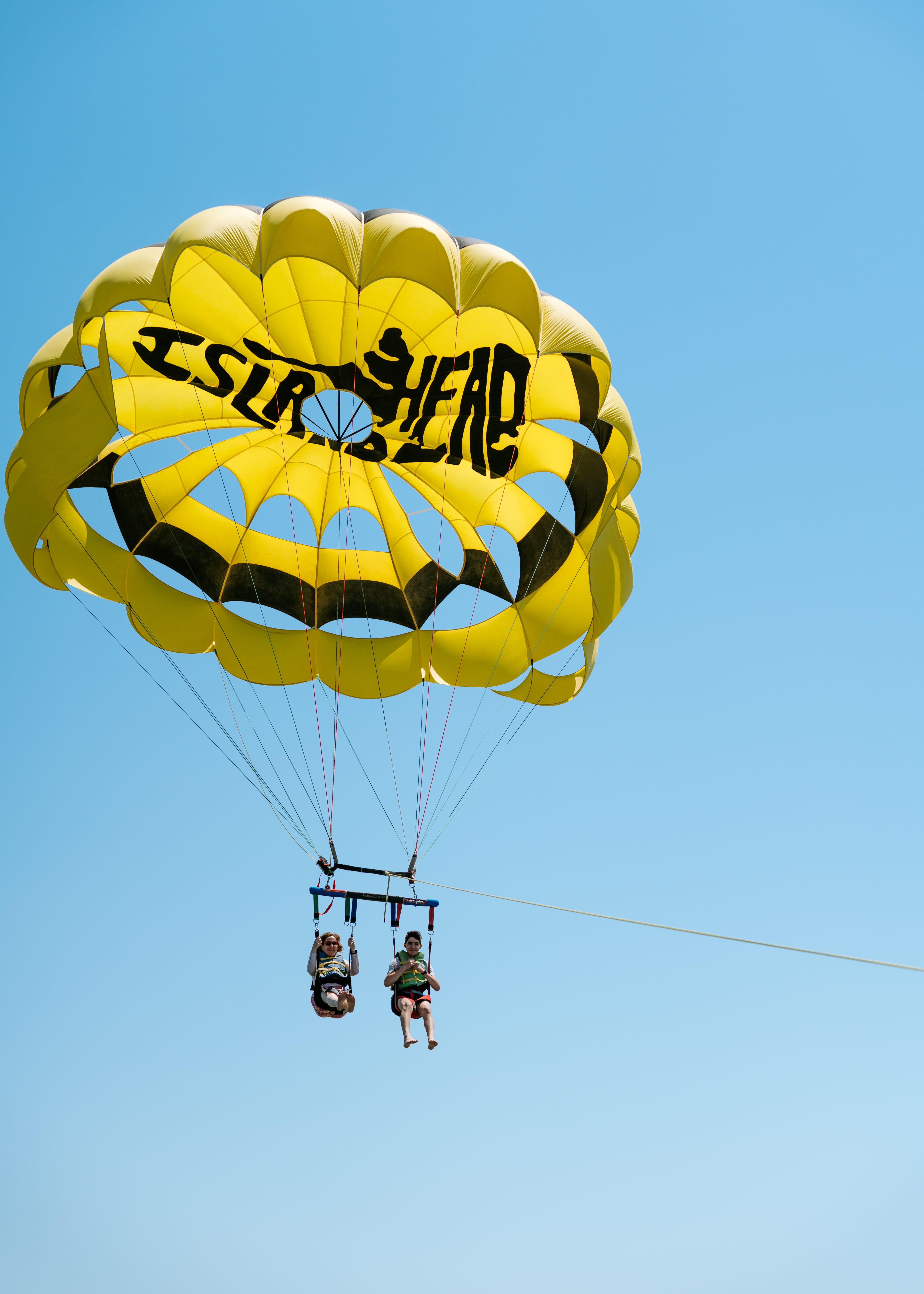 Two people tandem parasailing beneath a bright yellow-and-black canopy, suspended by harnesses and lines from a tow rope against a clear blue sky.