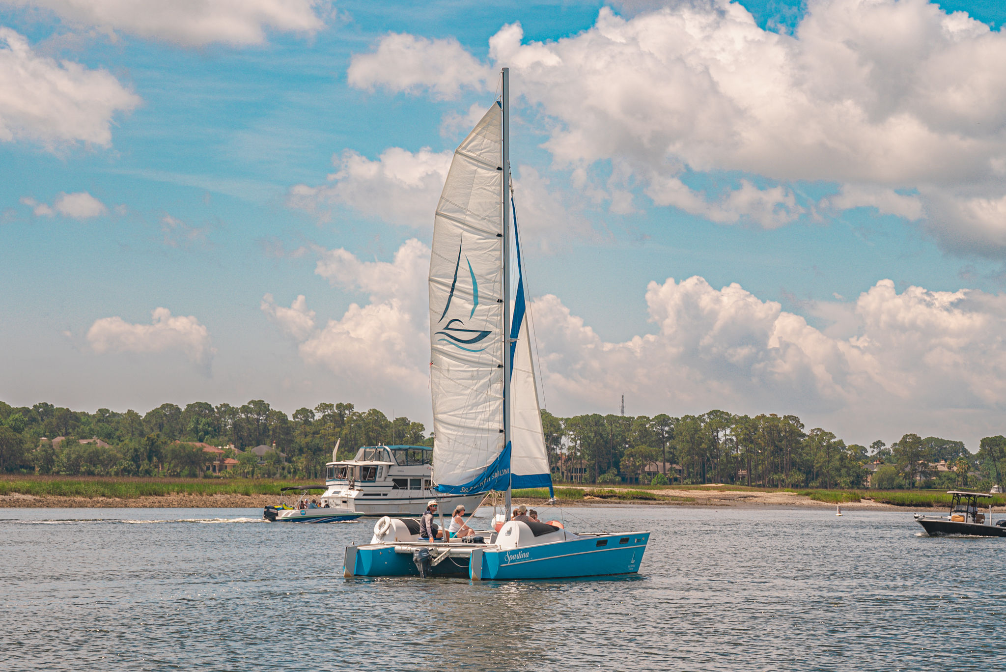 Blue catamaran sailboat with a tall white sail gliding on a calm coastal inlet, tree-lined shoreline and other motorboats under a bright sky with puffy clouds