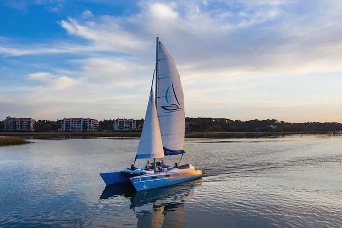 Catamaran sailboat with white sails gliding across a calm coastal inlet at sunset, blue sky with wispy clouds and waterfront condos reflected in the water.