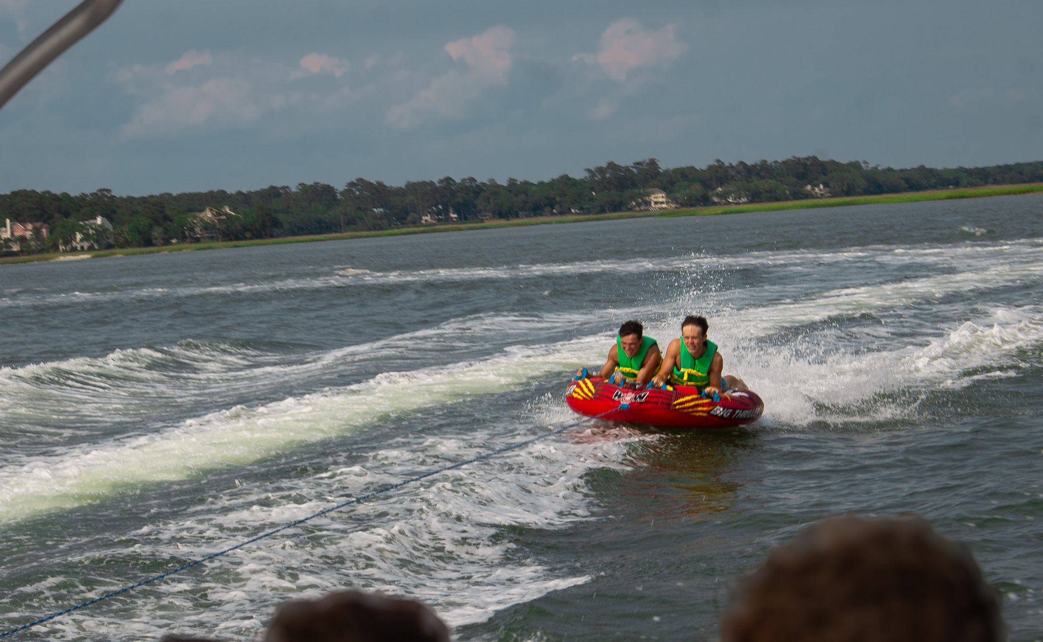 Two people wearing green life vests ride a bright red inflatable tube being towed across a coastal bay, splashing through the boat wake with a tree-lined shoreline and houses in the distance.