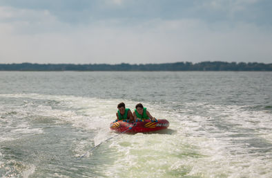 Two people in green life jackets ride a red inflatable towable, gripping handles as a boat pulls them across a lake through white wake under a partly cloudy sky