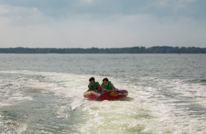 Two people in green life jackets ride a red inflatable towable, gripping handles as a boat pulls them across a lake through white wake under a partly cloudy sky