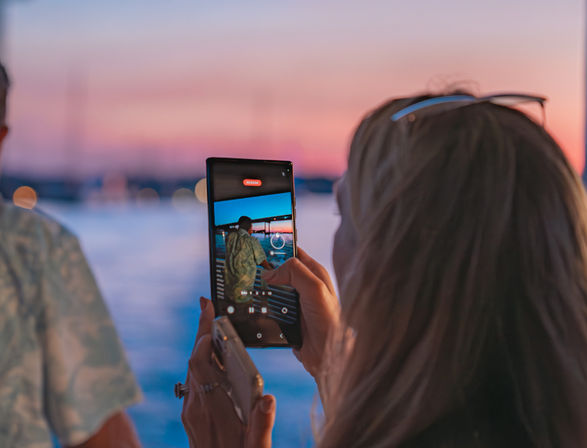 Person holding a smartphone recording a friend on a dock at a pastel-pink waterfront sunset, phone screen and marina bokeh visible