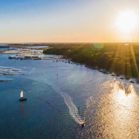 Aerial sunset over a coastal inlet with a white sailboat and a speedboat carving wakes past tree-lined docks and shimmering water.