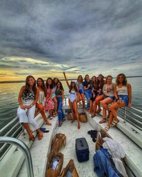Bachelorette boat party at sunset — group of friends on a boat bow, bride in a white "BRIDE" swimsuit raising her arm, calm water and dramatic cloudy sky.