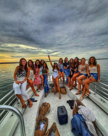 Bachelorette boat party at sunset — group of friends on a boat bow, bride in a white "BRIDE" swimsuit raising her arm, calm water and dramatic cloudy sky.