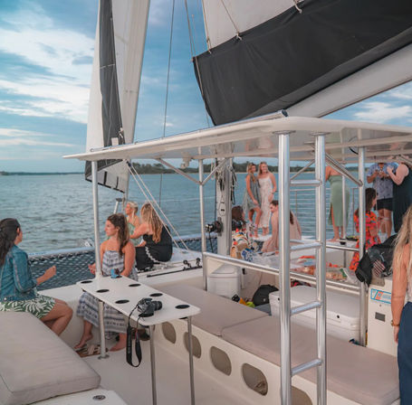 Group of people socializing on a sailing catamaran deck with raised sails, cushioned seating and coastal waters under a blue sky for a relaxed leisure cruise vibe.