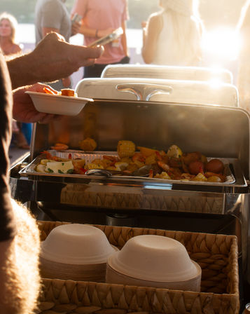 Sunset waterfront buffet with steaming seafood boil - shrimp, corn and potatoes in a chafing dish, guests serving into compostable bowls