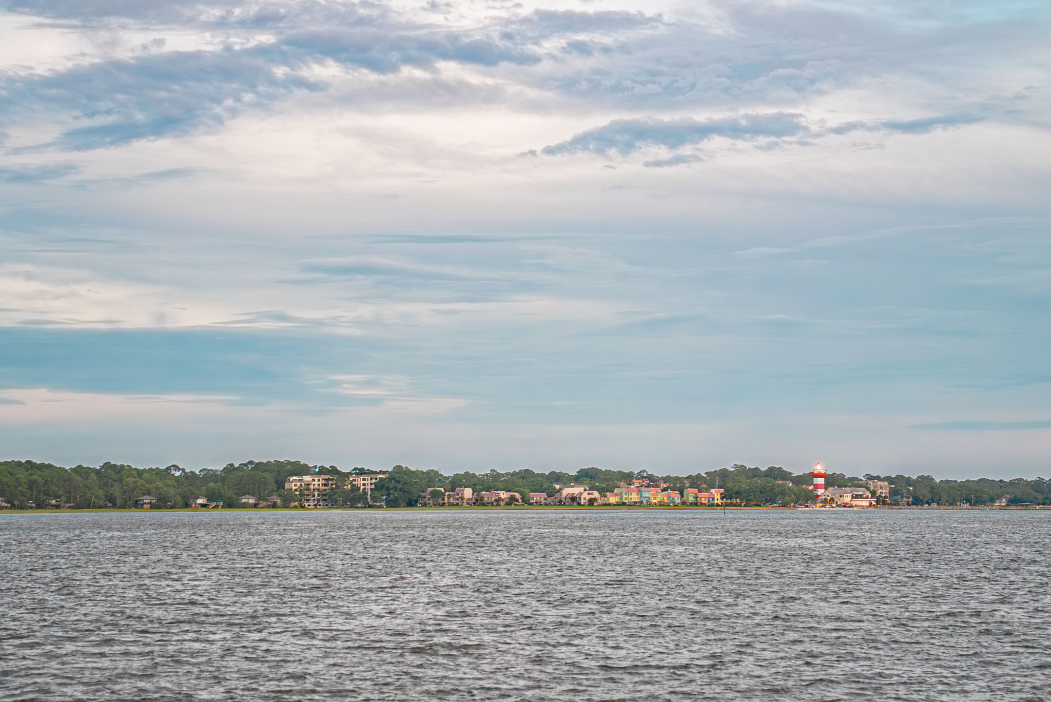 Panoramic bay view of a coastal town with a red-and-white lighthouse, colorful waterfront homes and tree-lined shore across calm water under a wide cloudy sky