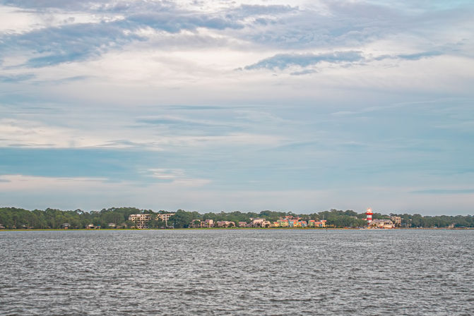 Panoramic bay view of a coastal town with a red-and-white lighthouse, colorful waterfront homes and tree-lined shore across calm water under a wide cloudy sky