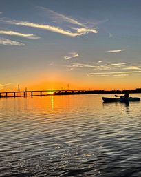 Silhouetted kayaker paddling on calm water at golden sunset, orange sky and clouds reflecting off a rippled river with a low bridge and power towers on the horizon.