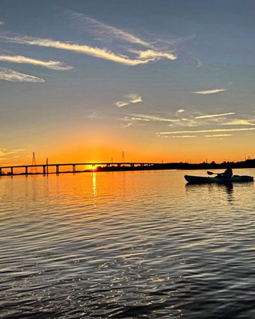 Silhouetted kayaker paddling on calm water at golden sunset, orange sky and clouds reflecting off a rippled river with a low bridge and power towers on the horizon.
