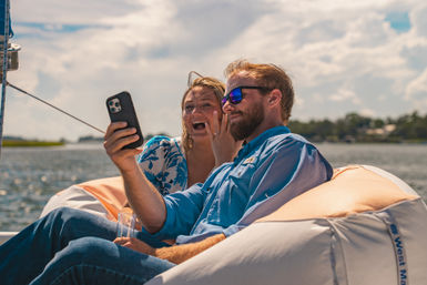 Couple laughing and taking a selfie on a sunny sailboat over calm coastal waters, man in sunglasses and woman showing a ring on her hand.
