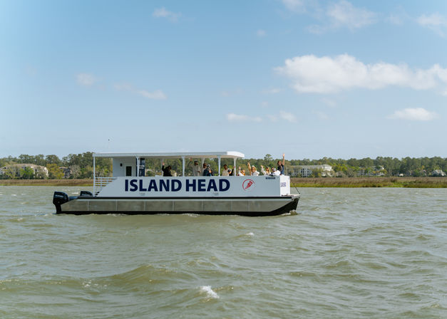White passenger boat with open deck and waving passengers cruising a coastal bay past marshy shoreline and waterfront homes under a clear blue sky