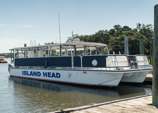 White catamaran-style passenger boat with navy side panels and large blue lettering docked at a sunny marina beside a wooden pier, calm water and tree-lined shore in the background.