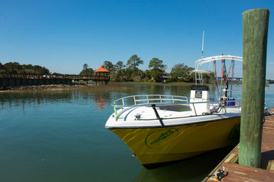 Bright yellow powerboat tied to a wooden dock by a tall green piling, reflected in calm coastal marsh water with a tree-lined boardwalk and gazebo under a clear blue sky