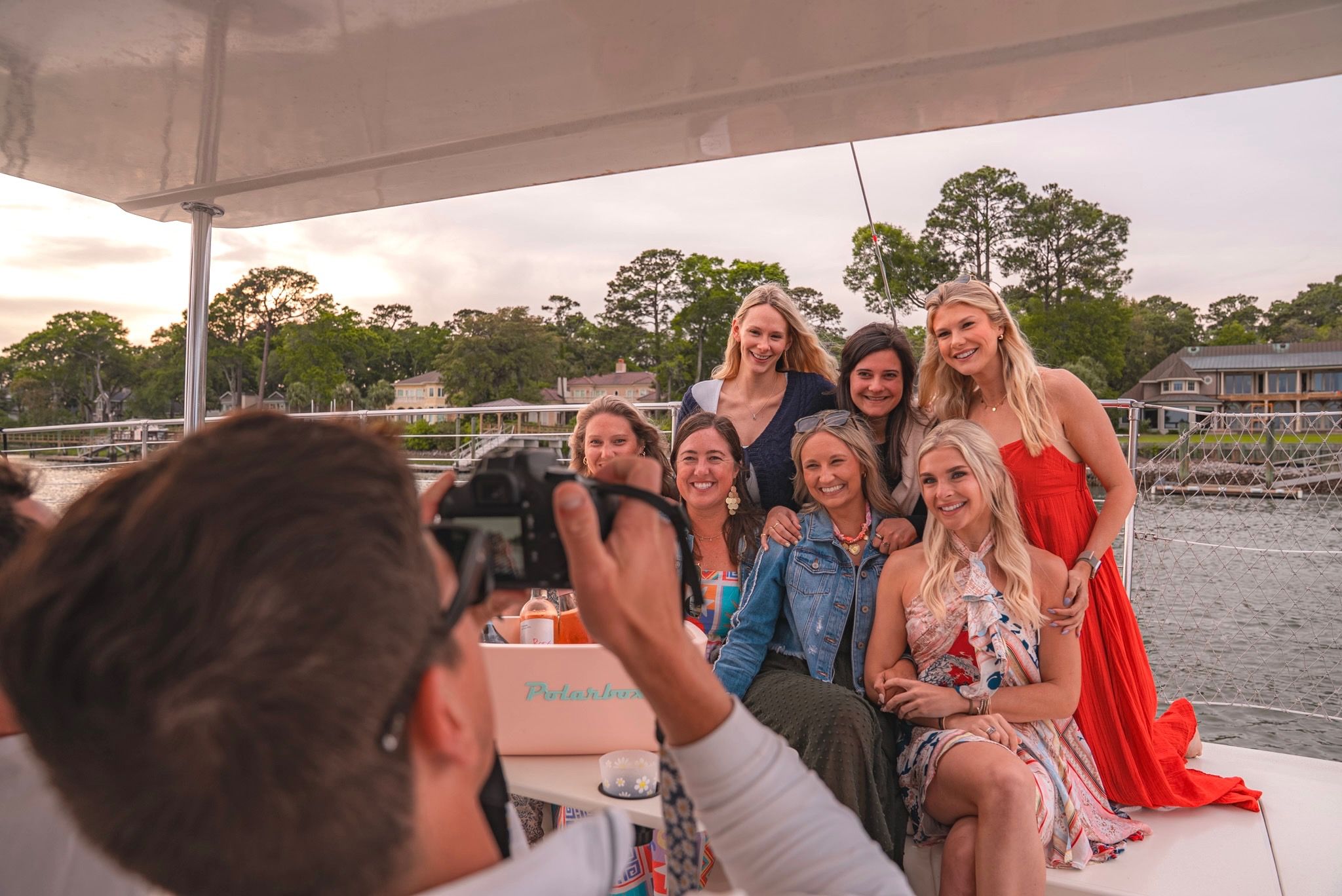 Photographer taking a group photo of seven women smiling on a sailboat near waterfront homes at sunset — relaxed coastal friends' outing.