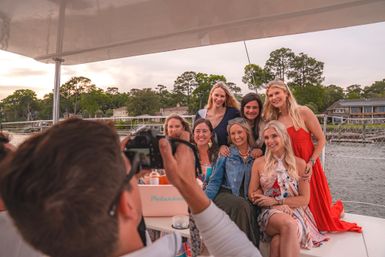 Photographer taking a group photo of seven women smiling on a sailboat near waterfront homes at sunset — relaxed coastal friends' outing.