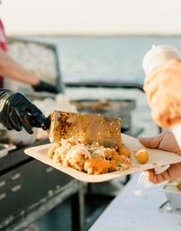Gloved cook using a spatula to plate saucy seafood rice and roasted potatoes onto a compostable tray at a waterfront food stall