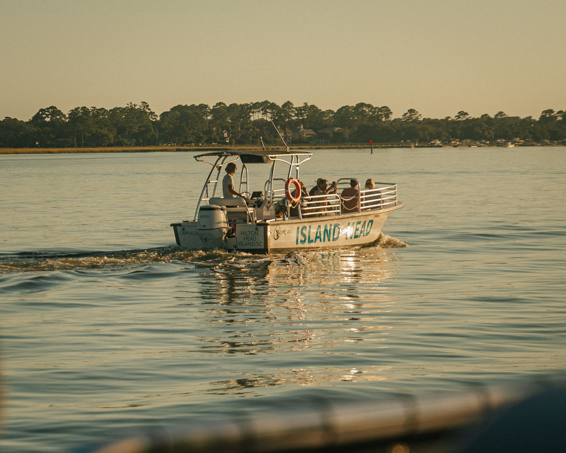 Small tour boat with passengers cruising calm waters at golden hour near Hilton Head Island, SC, with tree-lined shoreline and warm reflections on the water
