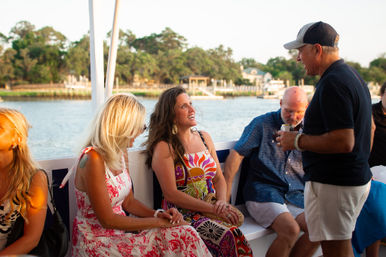 Friends chatting and laughing on a leisure boat at sunset, women in colorful summer dresses and men in casual shirts with a waterfront and trees in the background.