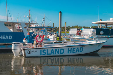 Sunny marina scene: white passenger boat with blue hull lettering docked at a coastal harbor, crew member at the helm, life ring on deck and clear reflection in calm water.