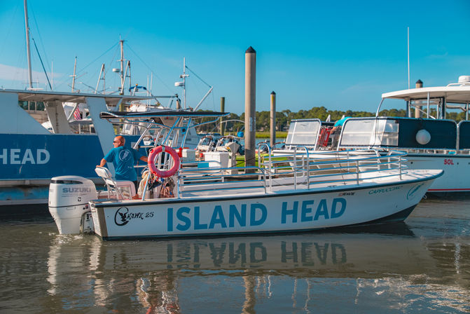 Sunny marina scene: white passenger boat with blue hull lettering docked at a coastal harbor, crew member at the helm, life ring on deck and clear reflection in calm water.