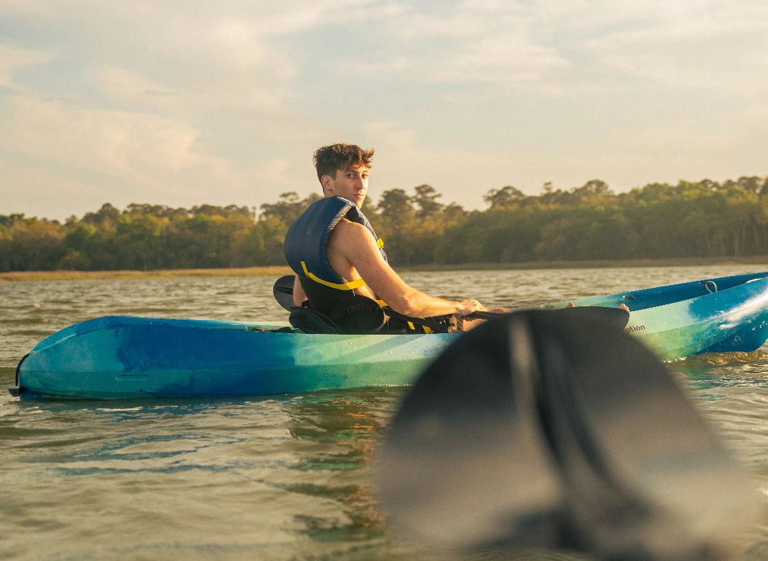 Solo kayaker in a blue kayak wearing a life jacket paddling on a calm lake at golden hour, paddle blade blurred in the foreground and tree-lined shore in the background